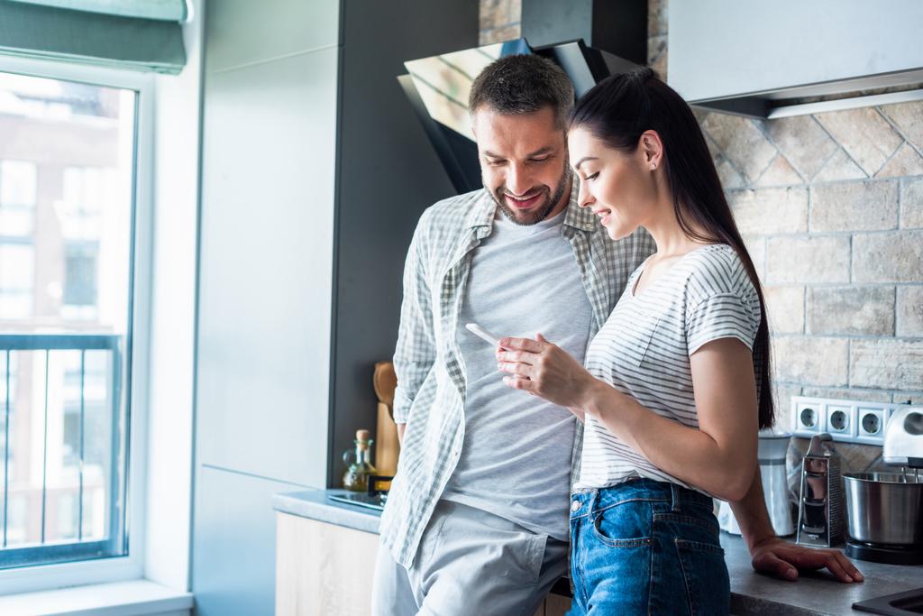 sonriente pareja usando teléfono inteligente juntos en la cocina, concepto de hogar inteligente
 - Foto, Imagen