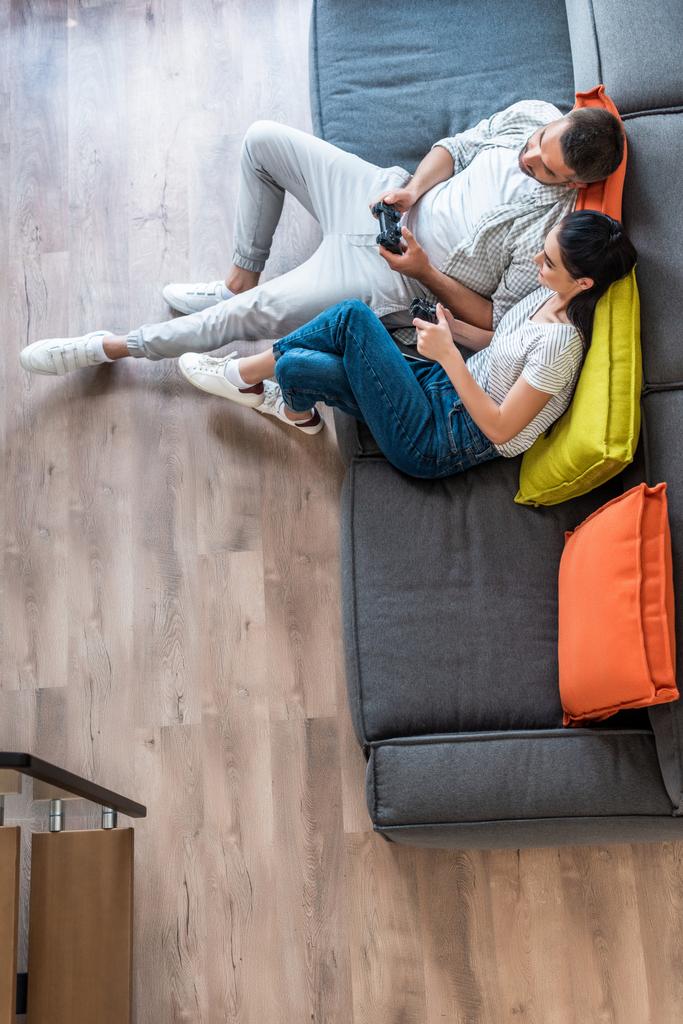 Overhead-Ansicht von Ehepaaren mit Gamepads, die gemeinsam Videospiele spielen, während sie zu Hause auf dem Sofa sitzen - Foto, Bild