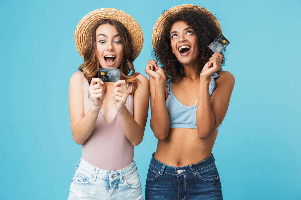 Foto dvou radostné multietnické žen 20s na sobě slaměné klobouky a letní oblečení, křičí, zatímco držení kreditky izolované modré pozadí - Fotografie, Obrázek