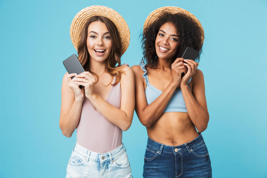 zwei reizende multiethnische Frauen 20er Jahre mit Strohhüten, die lächeln und beide ihr Smartphone in der Hand halten, während sie isoliert auf blauem Hintergrund in die Kamera schauen - Foto, Bild
