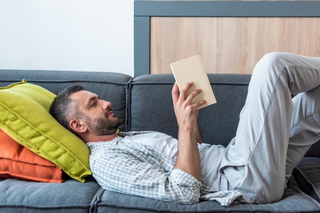 Widok z boku Brodaty mężczyzna czytanie książki leżąc na kanapie w domu - Zdjęcie, obraz