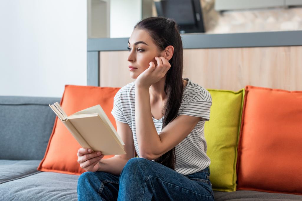 Porträt einer nachdenklichen Frau, die wegschaut, während sie zu Hause auf der Couch ein Buch liest - Foto, Bild