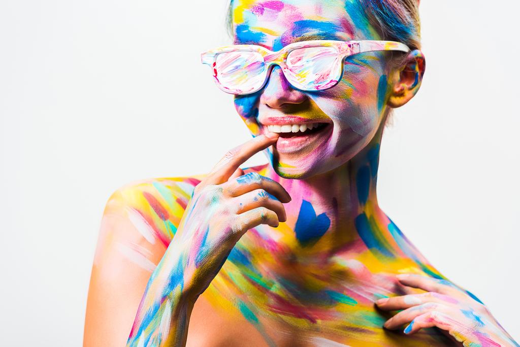 χαμογελώντας ελκυστική κοπέλα με πολύχρωμο φωτεινό σώμα τέχνη και γυαλιά ηλίου που αγγίζουν τα χείλη που απομονώνονται σε λευκό - Φωτογραφία, εικόνα