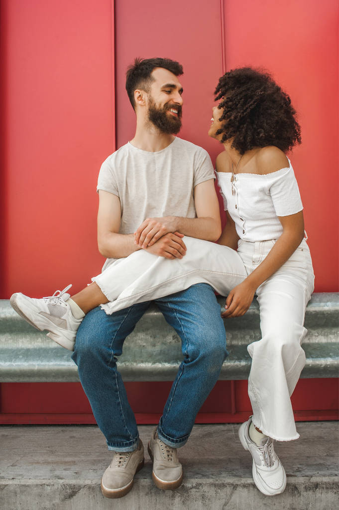 mooie gelukkige jonge sex tussen verschillendre rassen paar glimlachen elkaar zittend samen op straat - Foto, afbeelding