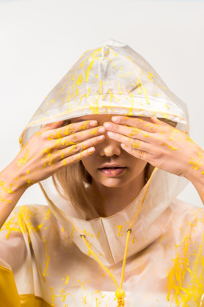 woman in raincoat painted with yellow paint covering eyes with hands isolated on white - Photo, Image