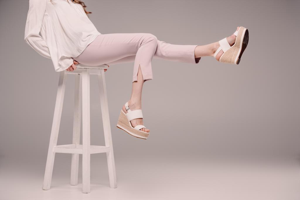 περικομμένη εικόνα της γυναίκας που παρουσιάζουν και δείχνουν τα πόδια στην καρέκλα σε γκρίζο φόντο  - Φωτογραφία, εικόνα