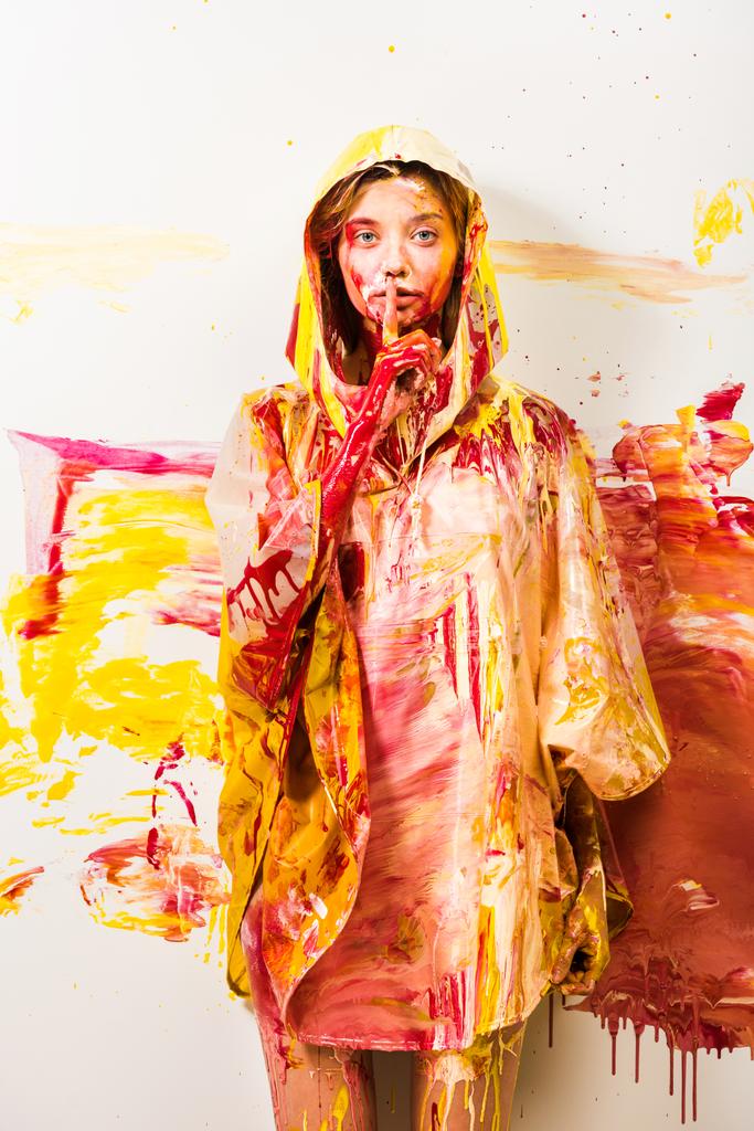 bela mulher em capa de chuva pintada com tintas amarelas e vermelhas mostrando gesto silêncio
 - Foto, Imagem