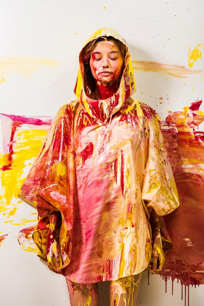 beautiful woman in raincoat painted with yellow and red paints standing with closed eyes - Photo, Image