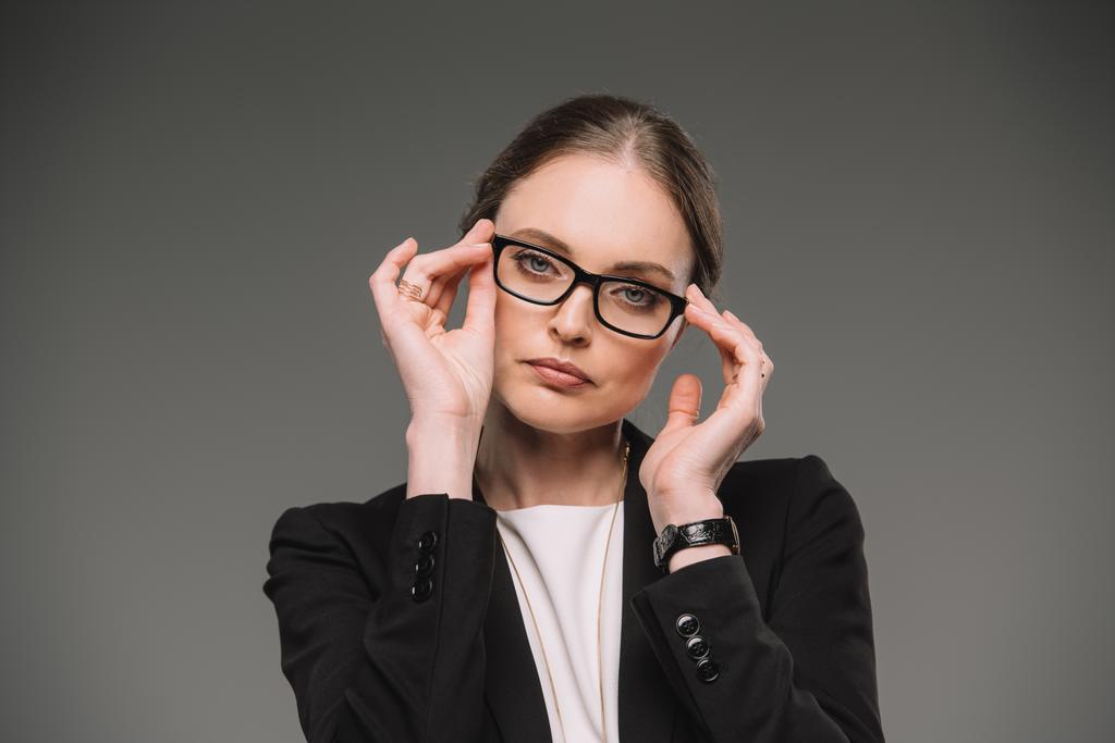 atractiva mujer de negocios ajustando gafas y mirando a la cámara aislada sobre fondo gris
  - Foto, imagen