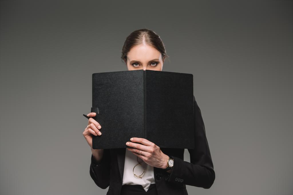 serious businesswoman covering face by textbook and looking at camera isolated on grey background  - Photo, Image