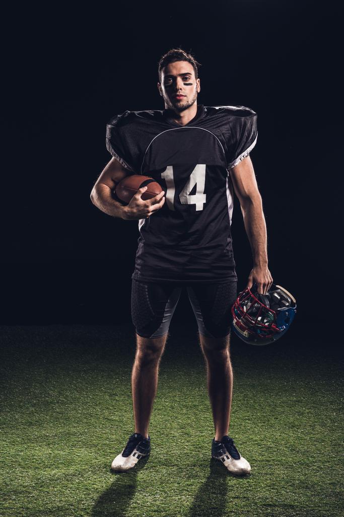 confiante jogador de futebol americano em uniforme preto segurando capacete e bola e olhando para a câmera em preto
 - Foto, Imagem