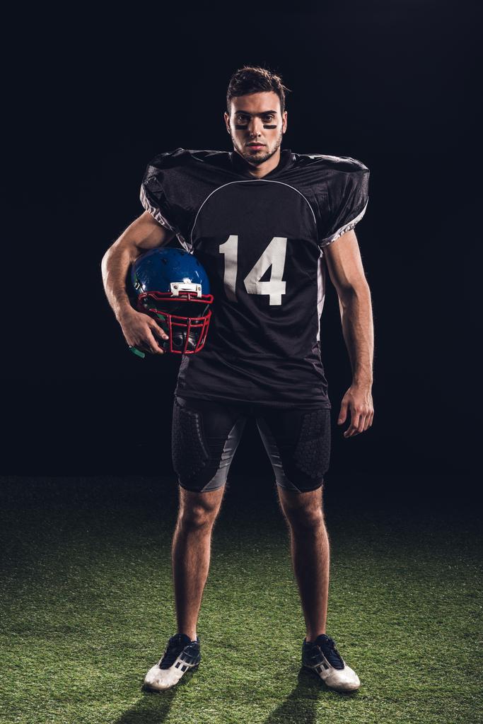 σοβαρή αμερικανικός παίκτης ποδοσφαίρου στην μαύρη στολή κρατώντας κράνος και κοιτάζοντας την κάμερα στο μαύρο - Φωτογραφία, εικόνα
