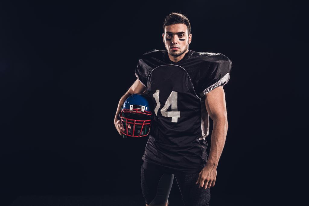 atletische american football speler in zwart uniform helm houden en kijken naar camera geïsoleerd op zwart - Foto, afbeelding