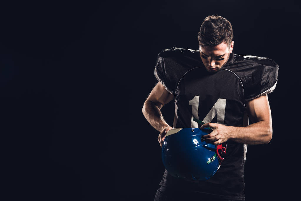 αμερικανικός παίκτης ποδοσφαίρου σε μαύρο χρώμα ομοιόμορφο με το κράνος στα χέρια που απομονώνονται σε μαύρο - Φωτογραφία, εικόνα