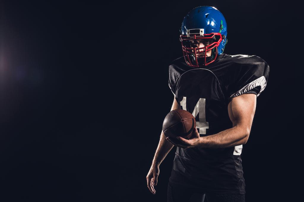 guapo jugador de fútbol americano con bola mirando a la cámara aislado en negro
 - Foto, imagen