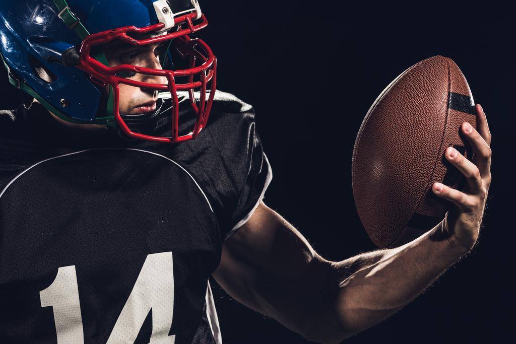Close-up πορτρέτο της εξοπλισμένη αμερικανικό ποδόσφαιρο παίκτης με μπάλα που απομονώνονται σε μαύρο - Φωτογραφία, εικόνα