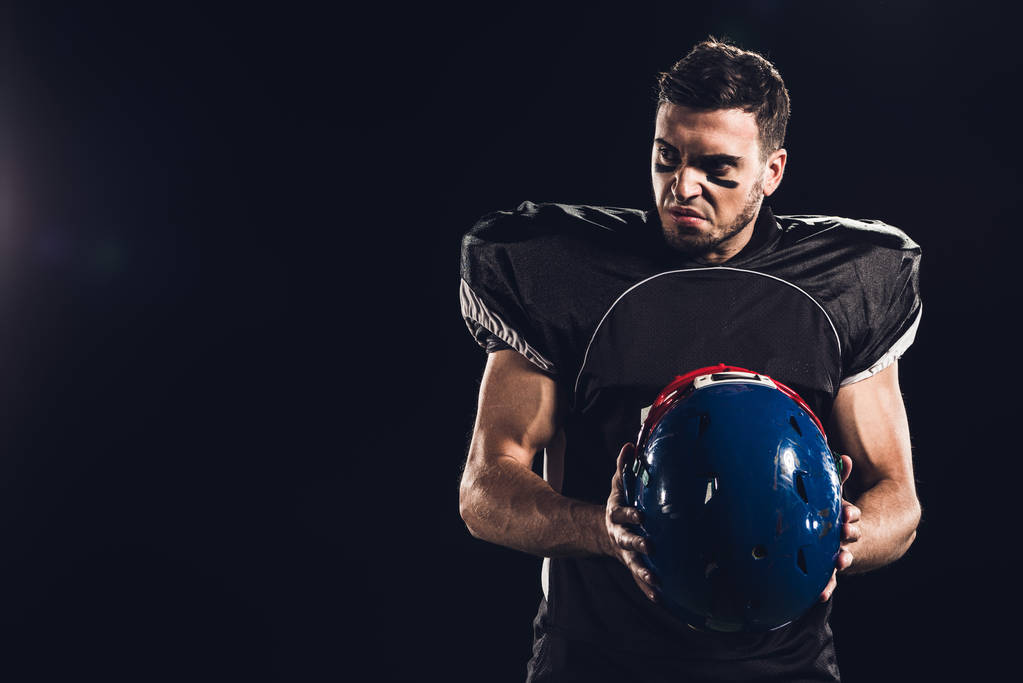 angry american football player in black uniform holding helmet and looking away isolated on black - Photo, Image