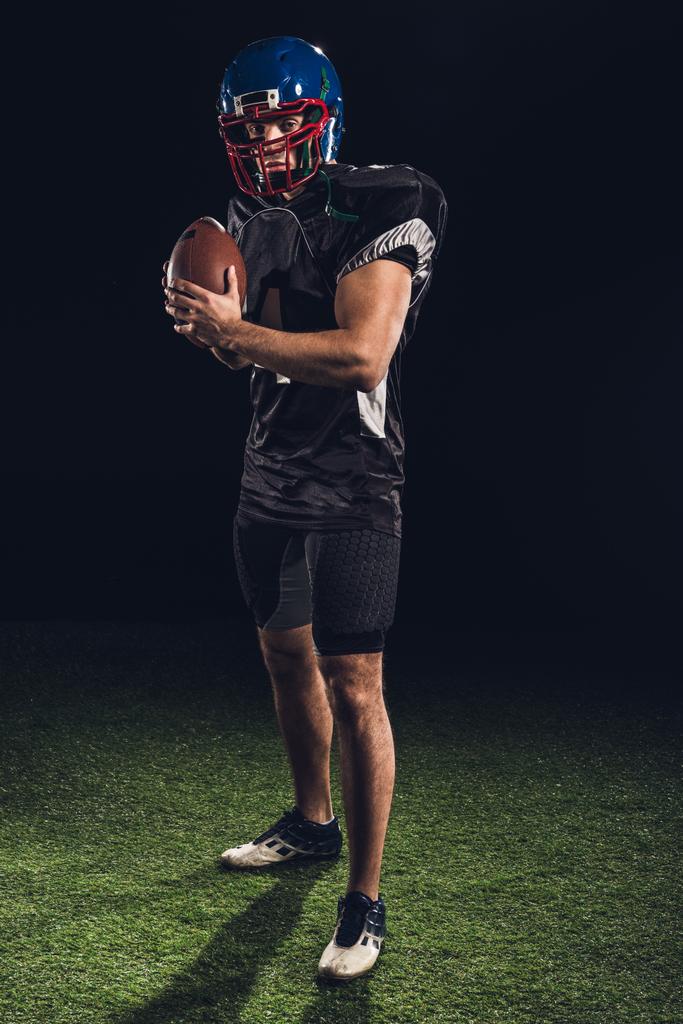 Американский футболист стоит на траве с мячом на черном и смотрит в камеру
 - Фото, изображение