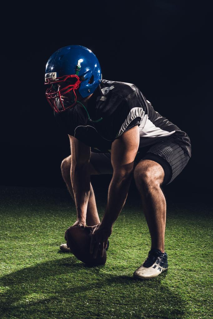 americký fotbalista stálý dřepy na trávě s míč mezi nohama na černém pozadí - Fotografie, Obrázek