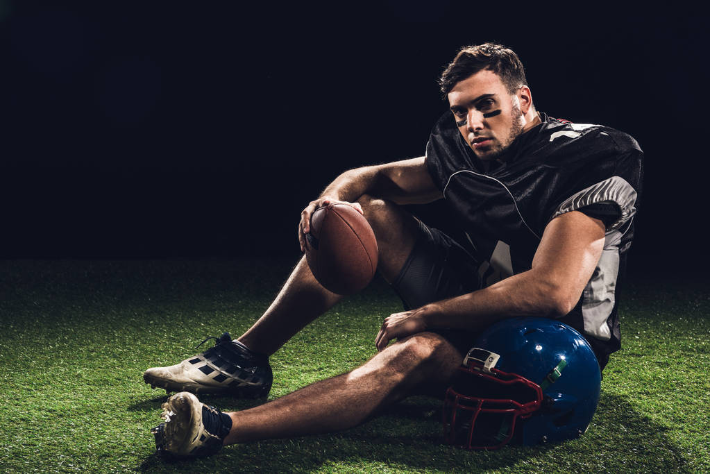 american football player sitting on grass with ball and helmet on black - Photo, Image