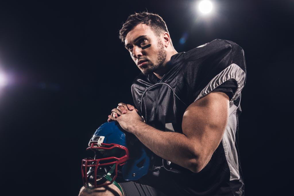 jogador de futebol americano com capacete olhando para longe sob holofotes em preto
 - Foto, Imagem