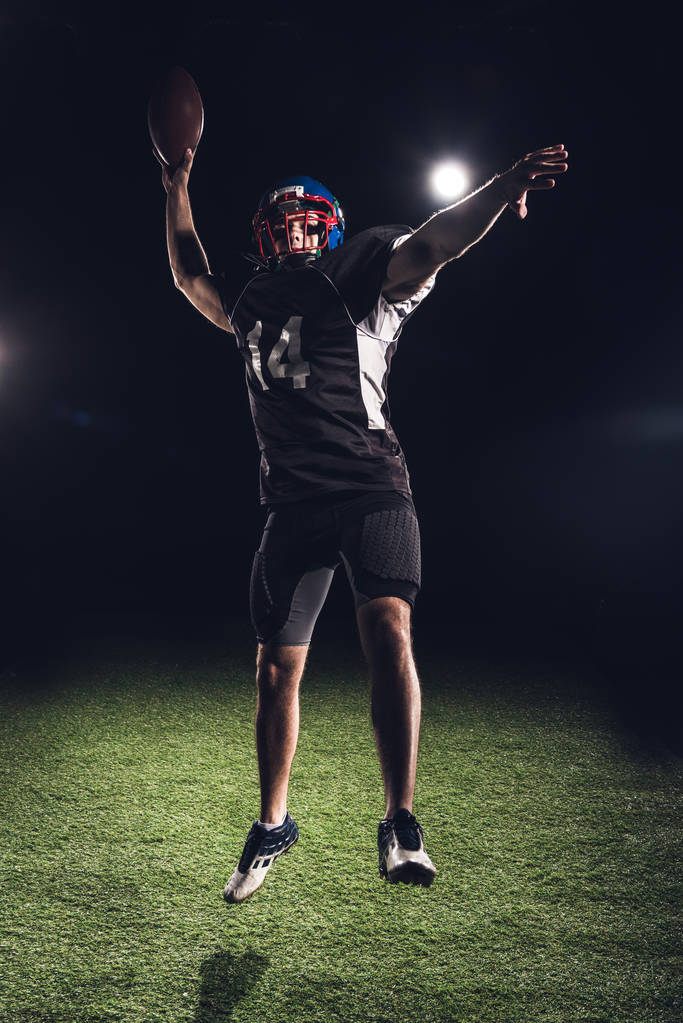athletischer American Football Spieler springt mit Ball unter Scheinwerfern auf schwarz - Foto, Bild