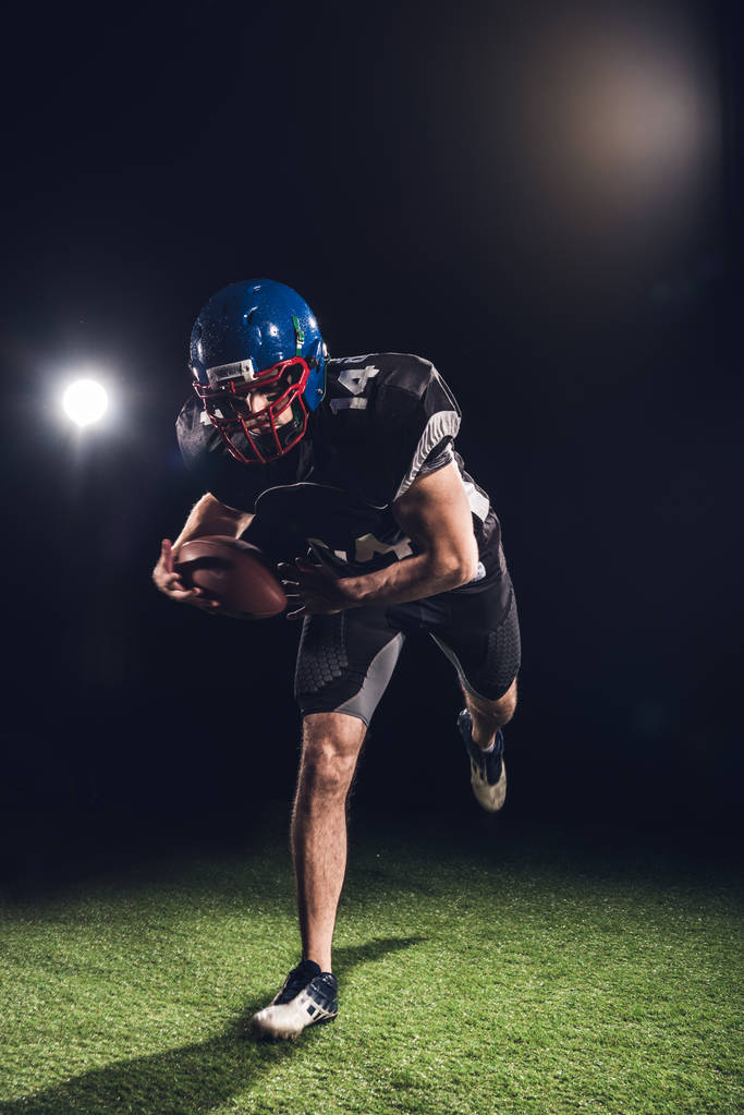 American Football-Spieler hält Ball und läuft auf dem Feld unter Scheinwerfern auf schwarz - Foto, Bild
