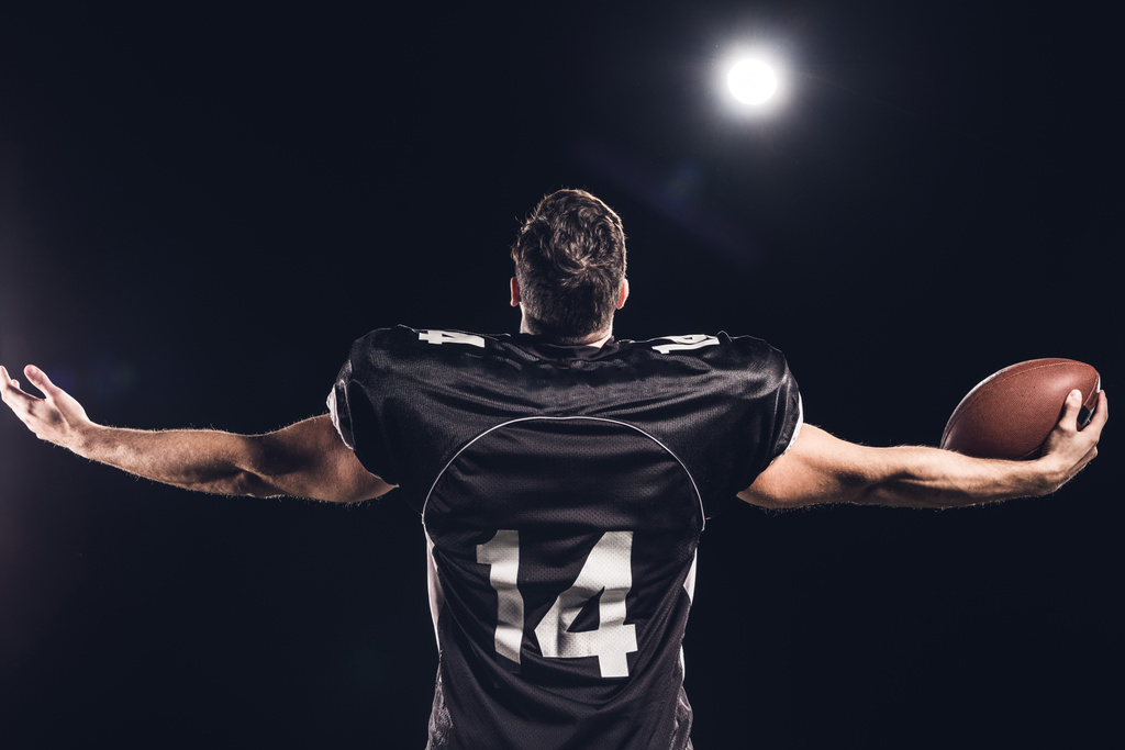 Rückansicht eines amerikanischen Fußballspielers mit Ball, der mit ausgestreckten Händen im Scheinwerferlicht auf schwarz nach oben schaut - Foto, Bild