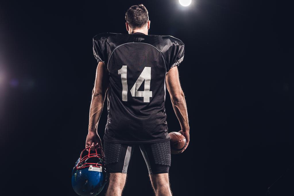 zadní pohled na hráče amerického fotbalu s míčem a helmu pod reflektor na černém pozadí - Fotografie, Obrázek