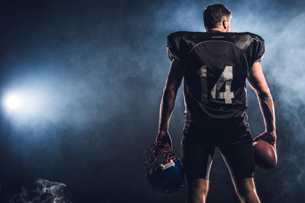 vista trasera del jugador de fútbol americano equipado con casco y bola en las manos contra el humo blanco
 - Foto, imagen