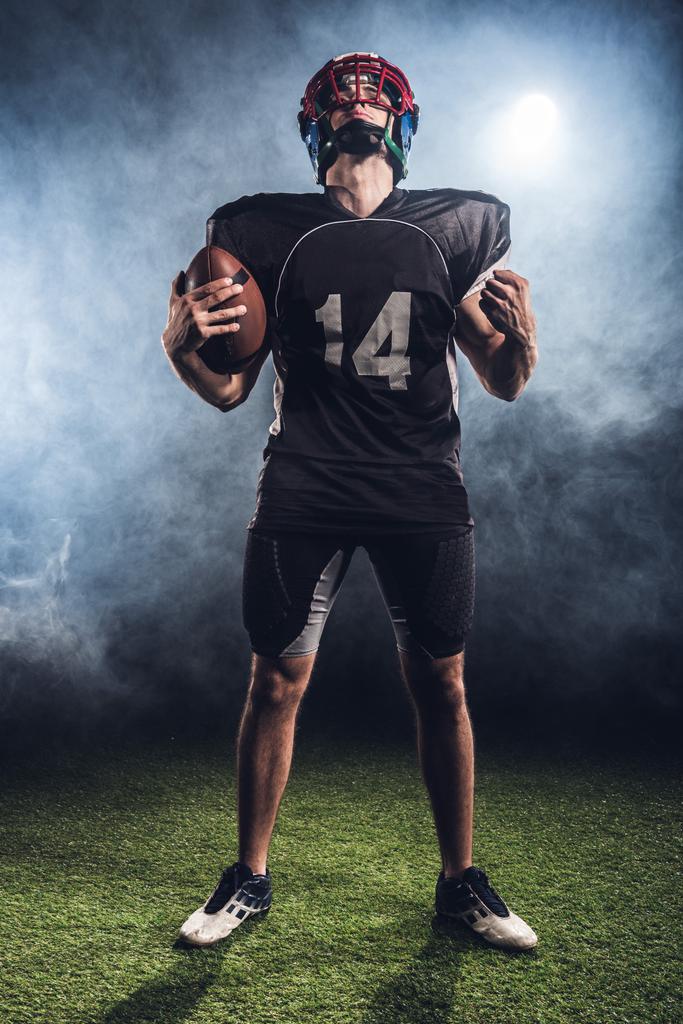 αμερικανικό ποδόσφαιρο παίκτης με μπάλα κάνοντας γροθιά και ψάχνει αντιμέτωποι με λευκό καπνό - Φωτογραφία, εικόνα