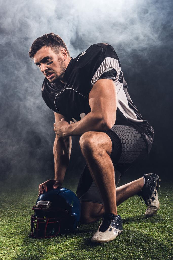 θυμωμένος νεαρός ποδοσφαιριστής Αμερικανός με κράνος στέκεται στο γόνατο σε μαύρο - Φωτογραφία, εικόνα