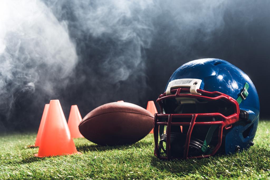 close-up shot of american football helmet with cones and ball on green grass with white smoke above - Photo, Image