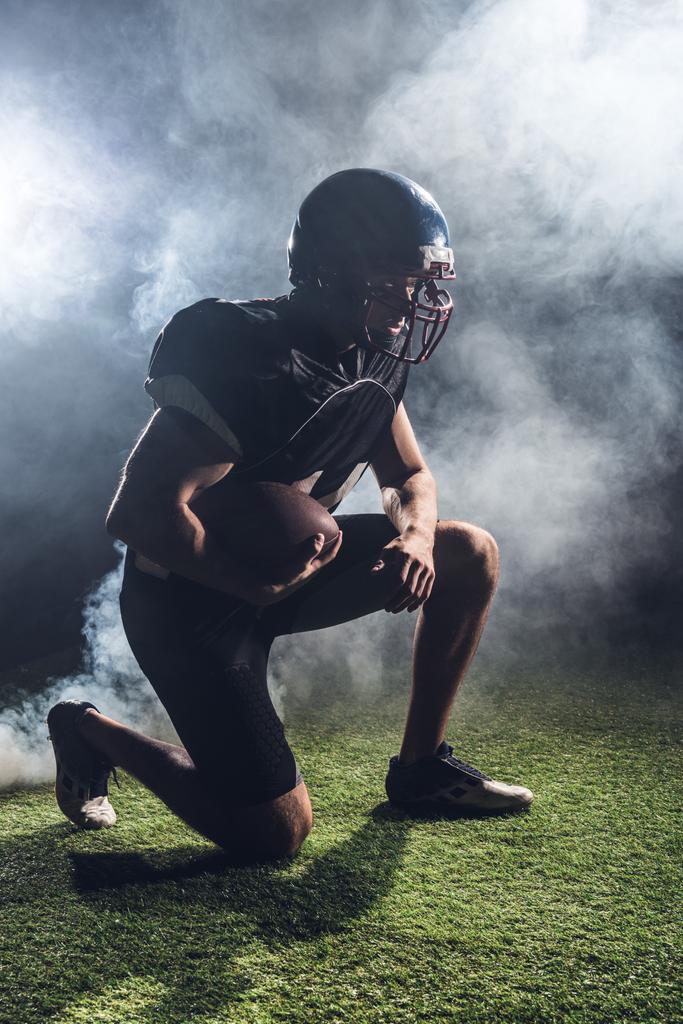 白い煙に対して手にボールで緑の草に膝の上に立って若いアメリカン ・ フットボール プレーヤー - 写真・画像
