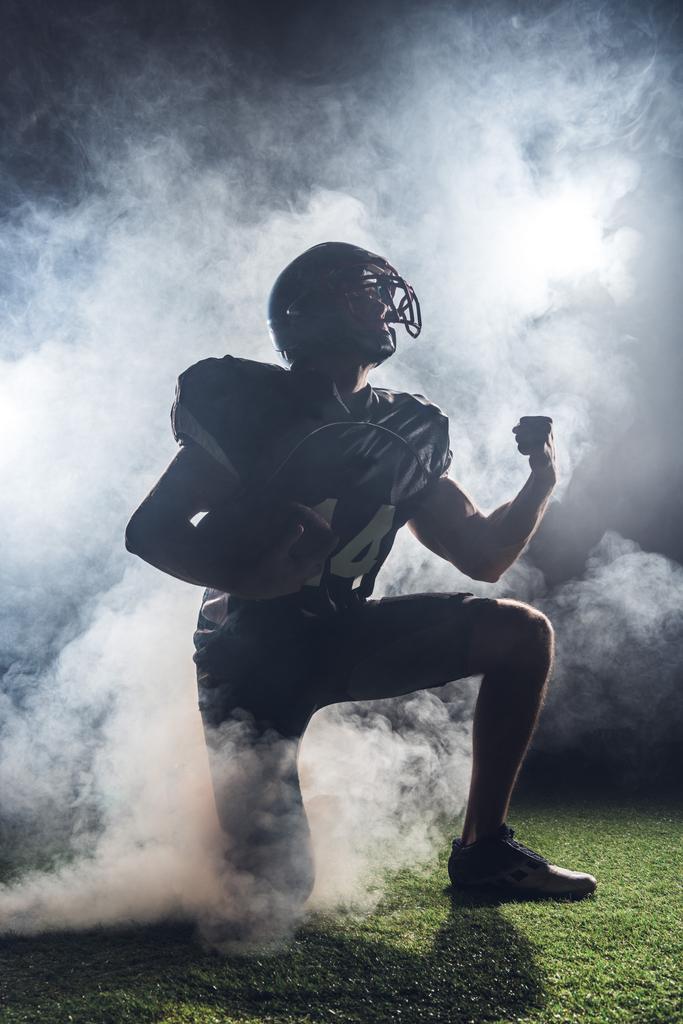jonge american football speler permanent op knie op gras en het verhogen van de vuist in witte rook - Foto, afbeelding