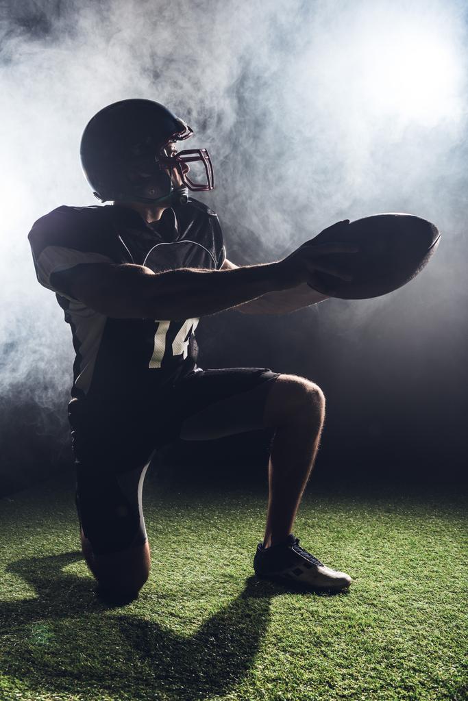 jonge american football speler permanent op knie op groene gras en houden bal tegen witte rook - Foto, afbeelding