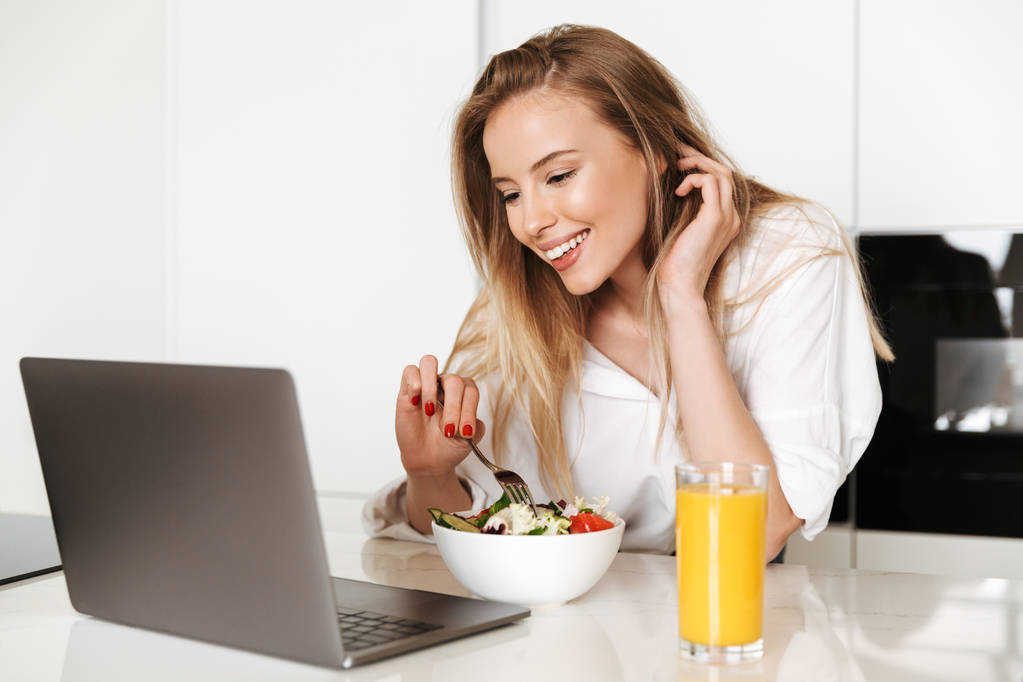 Šťastná mladá žena jíst salát z mísy a pít pomerančový džus stoje na kuchyň a sledování filmu na laptop - Fotografie, Obrázek