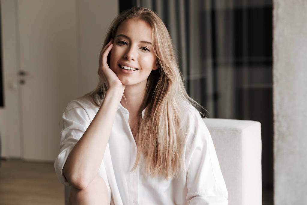 Прекрасна молода жінка, одягнена в білу сорочку, сидить на дивані вдома і дивиться на камеру
 - Фото, зображення