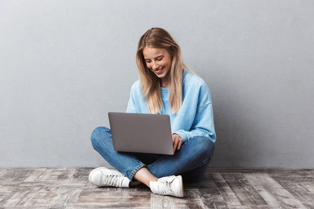 Menina loira feliz usando computador portátil enquanto sentado com as pernas cruzadas isolado sobre fundo cinza
 - Foto, Imagem