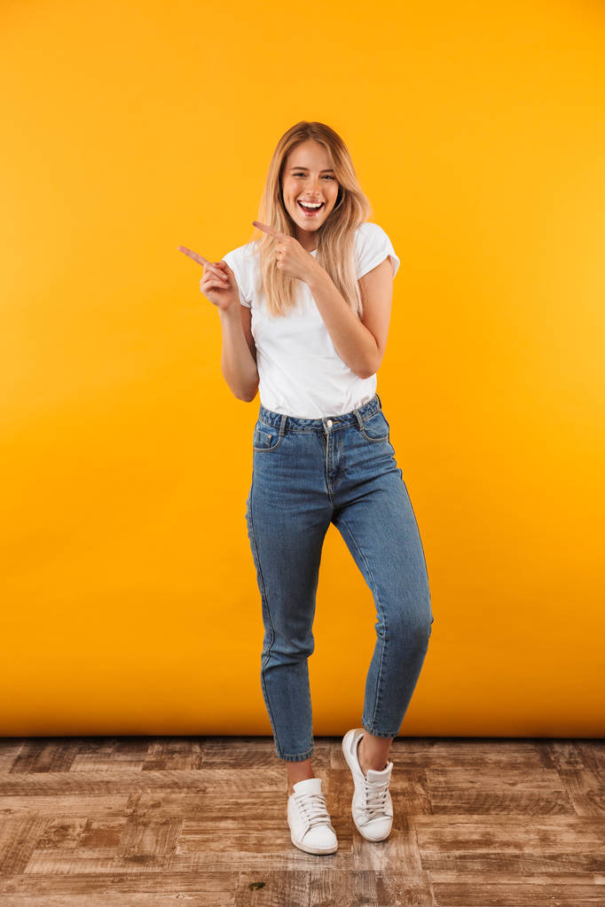 Retrato de comprimento total de uma jovem loira alegre apontando para o espaço de cópia isolado sobre fundo amarelo
 - Foto, Imagem