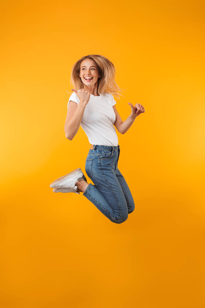Retrato de larga duración de una alegre joven rubia apuntando hacia afuera mientras salta aislada sobre un fondo amarillo
 - Foto, Imagen