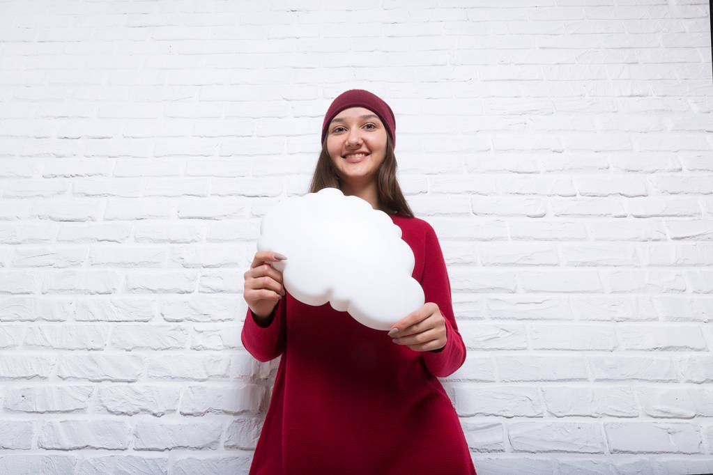 A young pretty smiling girl in a red sweater and a hat against the background of a modern white brick wall in the loft style holds white clouds in her hands. She looks at the viewer. Copy space. - Photo, Image