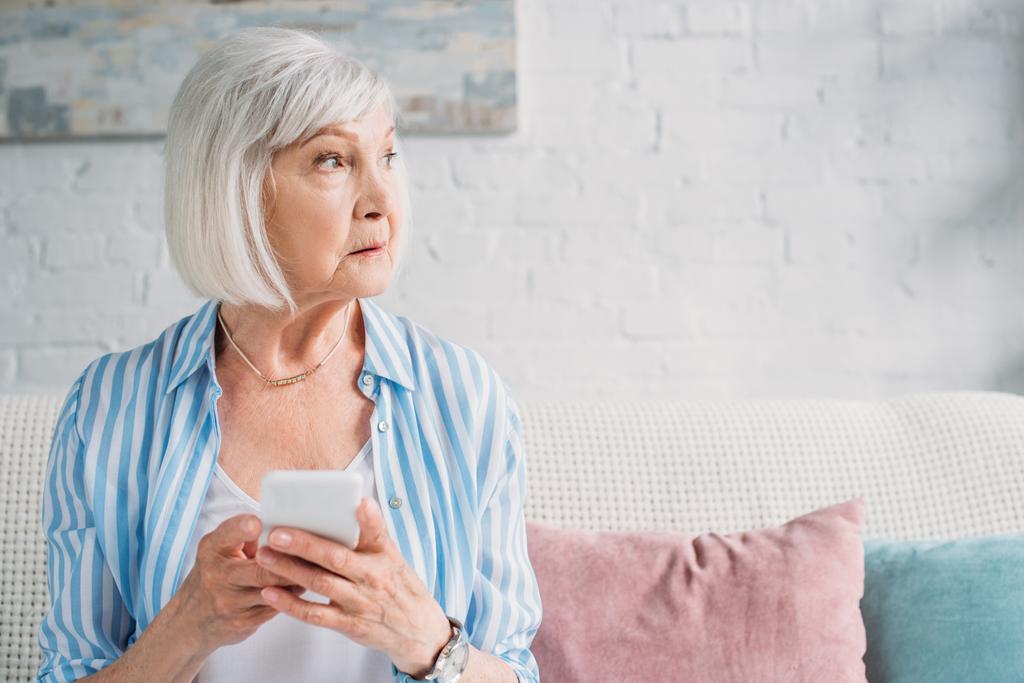 Porträt einer nachdenklichen Seniorin mit Smartphone, die auf dem heimischen Sofa wegschaut - Foto, Bild