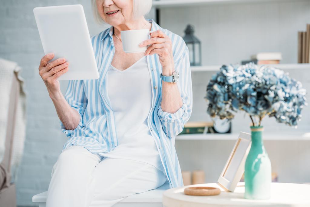Teilansicht einer Seniorin mit Tasse Kaffee zu Hause mit Tablet - Foto, Bild