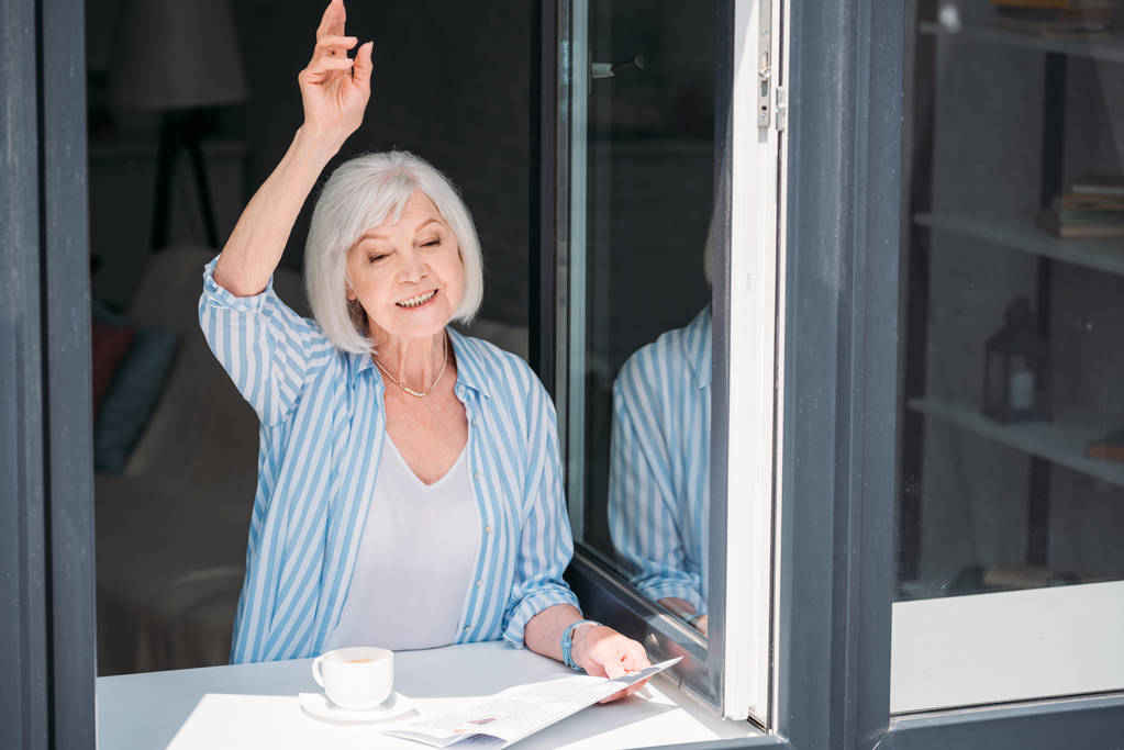mujer mayor saludando a alguien mientras está de pie en el alféizar de la ventana con periódico y taza de café aromático en casa
 - Foto, Imagen