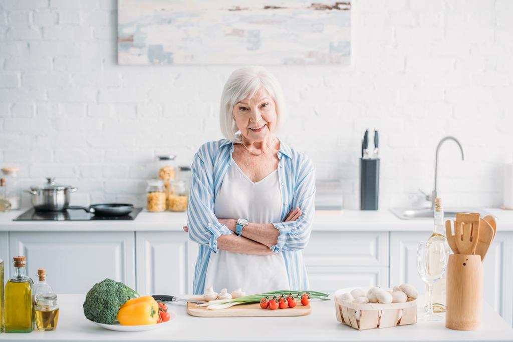 Portret van lachende senior met armen gekruist staande bij de balie met verse groenten in de keuken - Foto, afbeelding