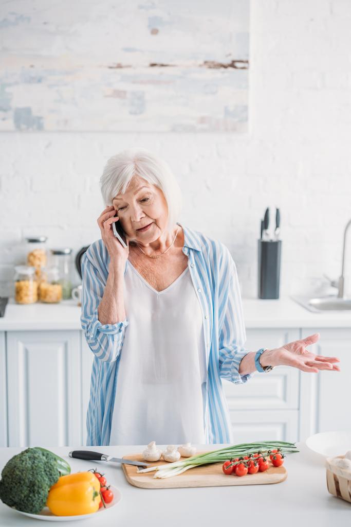 portrét starší ženy mluví o smartphone stoje u pultu s čerstvou zeleninou na prkénku v kuchyni - Fotografie, Obrázek
