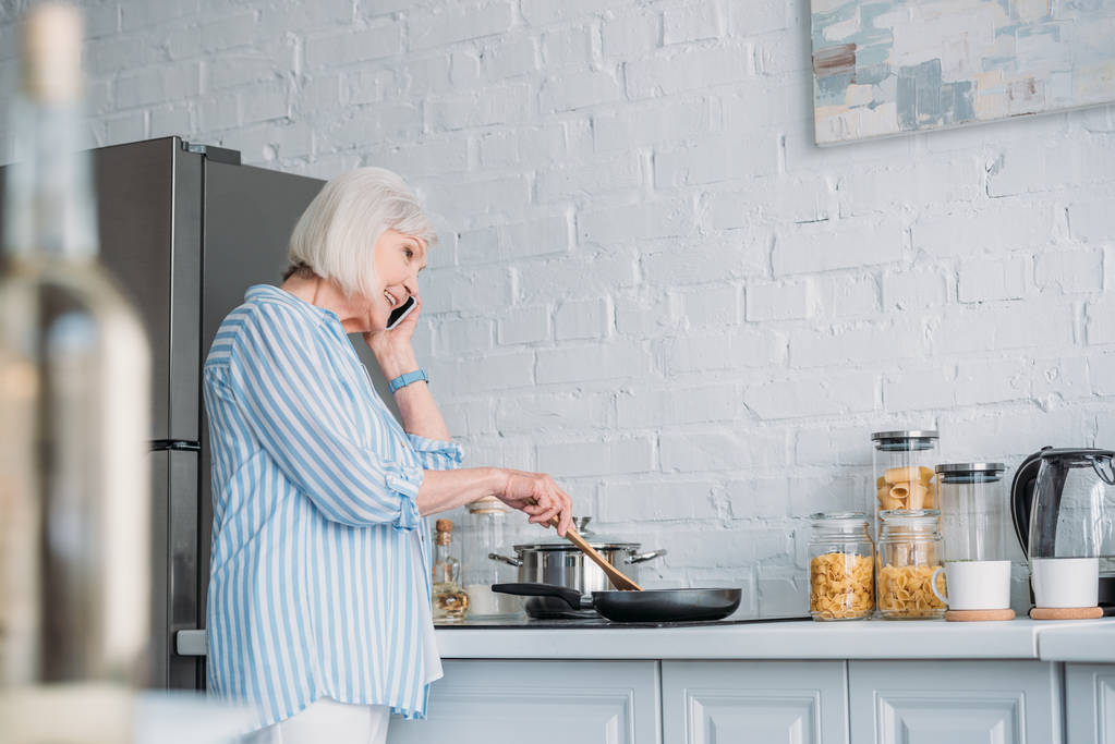 Widok z boku uśmiechający się starszy kobieta rozmawia na smartfonie podczas gotowanie w kuchni - Zdjęcie, obraz