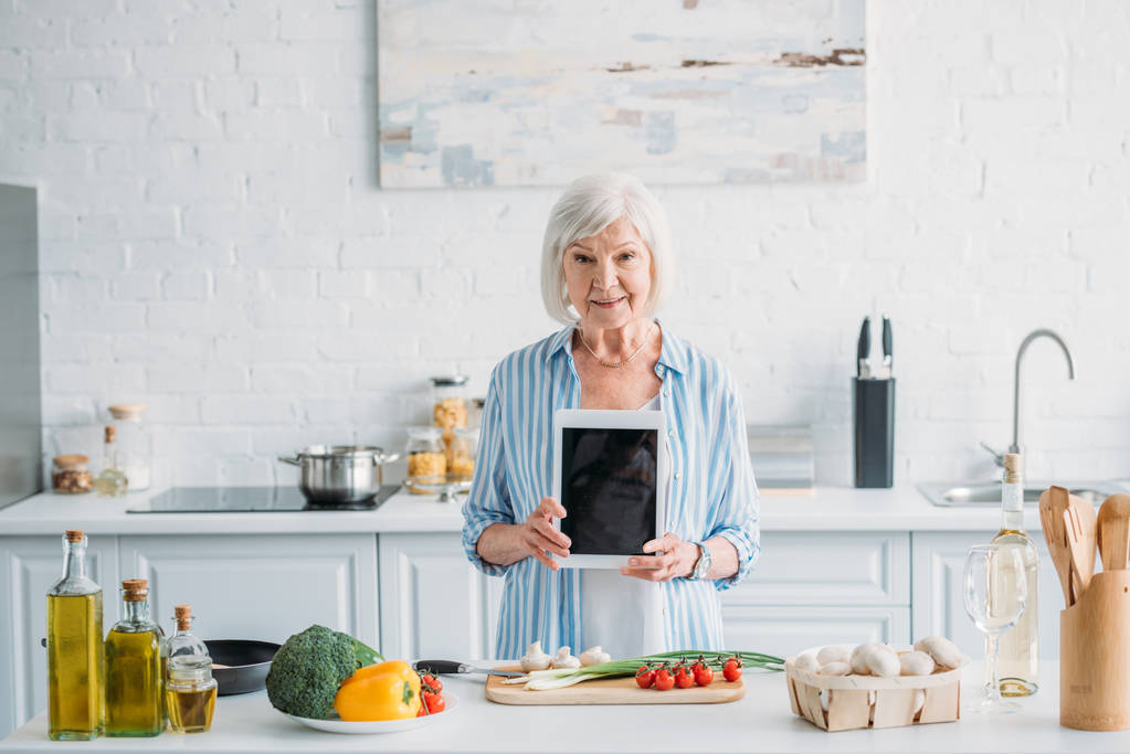 キッチンで新鮮な野菜とカウンターに立っている空白の画面と年配の女性示すタブレットの肖像画 - 写真・画像