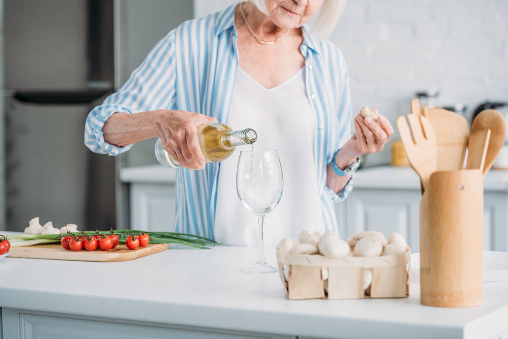 gedeeltelijke weergave van senior vrouw gieten van wijn in glazen aan balie met verse groenten in de keuken - Foto, afbeelding